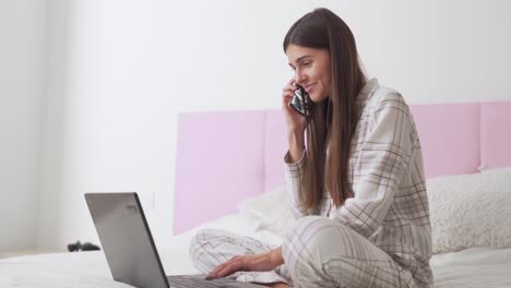linda chica en pijama en la cama hablando por teléfono y trabajando en una laptop