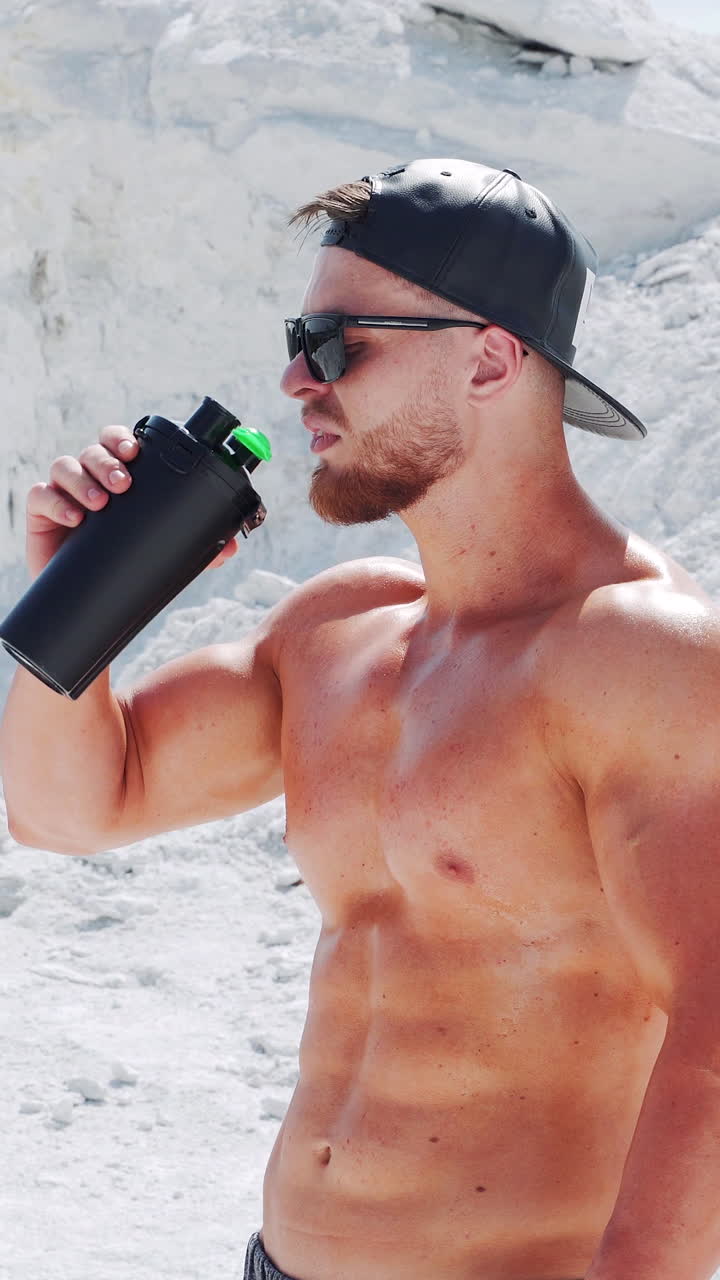 Strong shirtless athlete drinks water from a bottle. Training in sports in nature. Vertical video