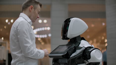 a man in a shirt communicates with a white robot asking questions and pressing the screen with his fingers