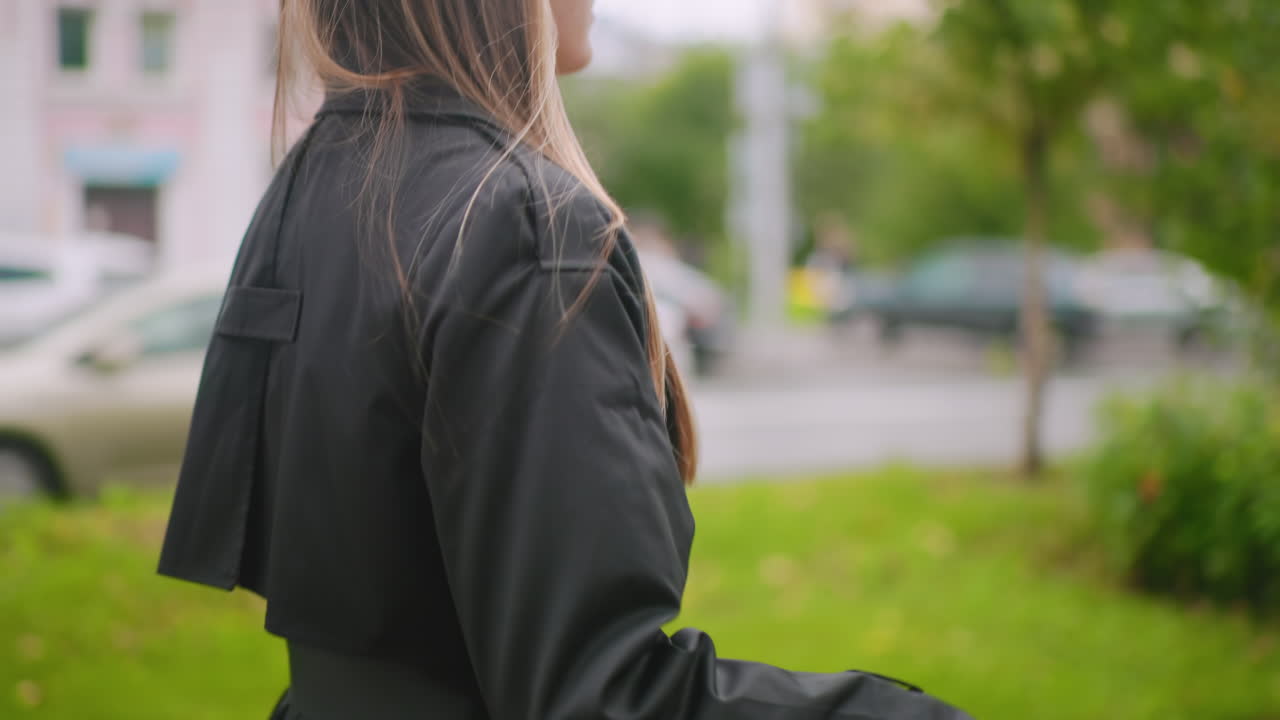 Close up of woman in black trench coat walking on colorful pavement holding closed umbrella with wooden handle, stylish footwear visible, urban street background with parked cars and greenery in autumn