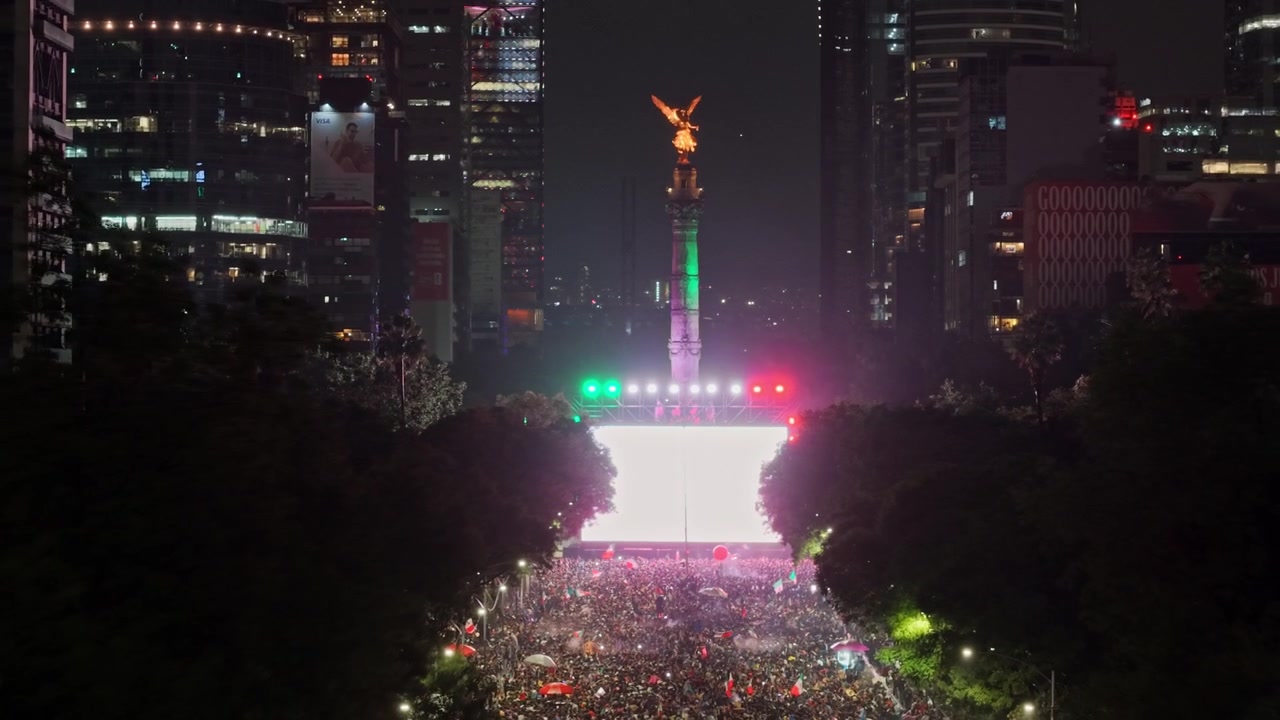 Vista aerea con teleobiettivo del Monumento all'Indipendenza con schermi e luci colorate di notte, che mostra persone che celebrano la vittoria del Messico alla Coppa del Mondo.