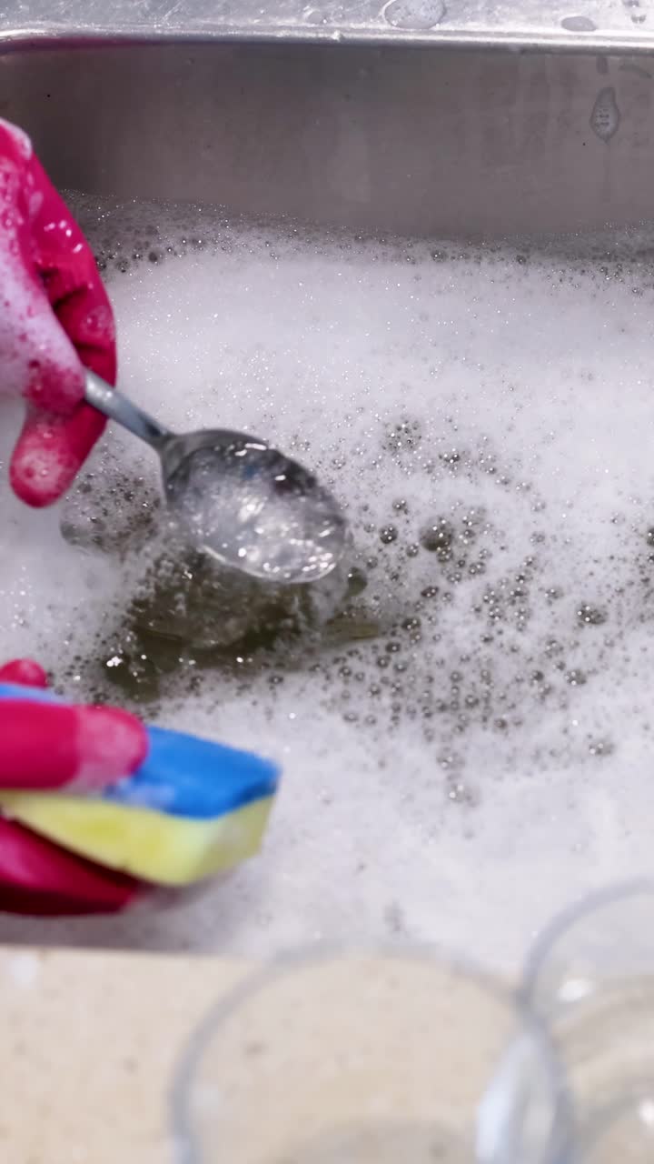 Una persona con i guanti pulisce un cucchiaio con una spugna in un lavello da cucina schiumoso.
