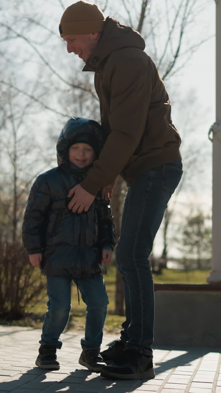 un primer plano de un padre levantando a su hijo ambos están vestidos con vaqueros y botas, con el niño con una chaqueta hinchada y el hombre con un gorro, árboles y un poste eléctrico están alrededor