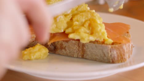 scrambled eggs being gently placed on top of smoked salmon on rustic sourdough bread in kitchen. close up breakfast food photography.