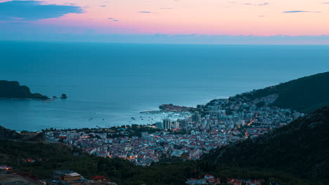 Day-to-night-time-lapse-of-the-seaside-city-Budva,-Montenegro-starting-with-a-glowing-orange-sky-and-transitioning-to-a-dark-sky-lit-by-the-glow-of-the-city