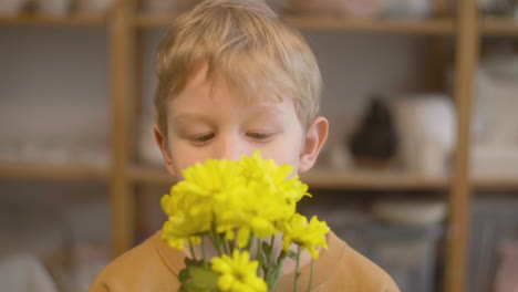 vista ravvicinata di un bambino biondo che sente l'odore di fiori gialli e guarda la macchina fotografica in un laboratorio artigianale