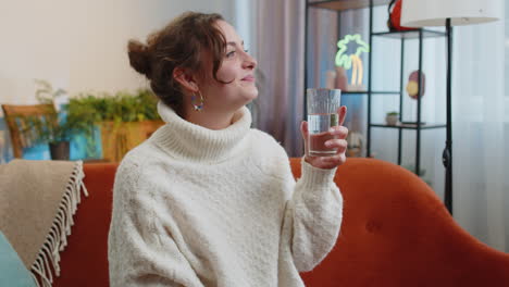 Thirsty-young-woman-sitting-at-home-holding-glass-of-natural-aqua-make-sips-drinking-still-water