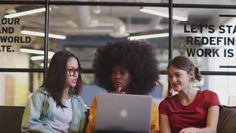 Three-millennial-women-in-a-casual-meeting-in-an-office-lounge-using-a-laptop-together,-tilt-shot