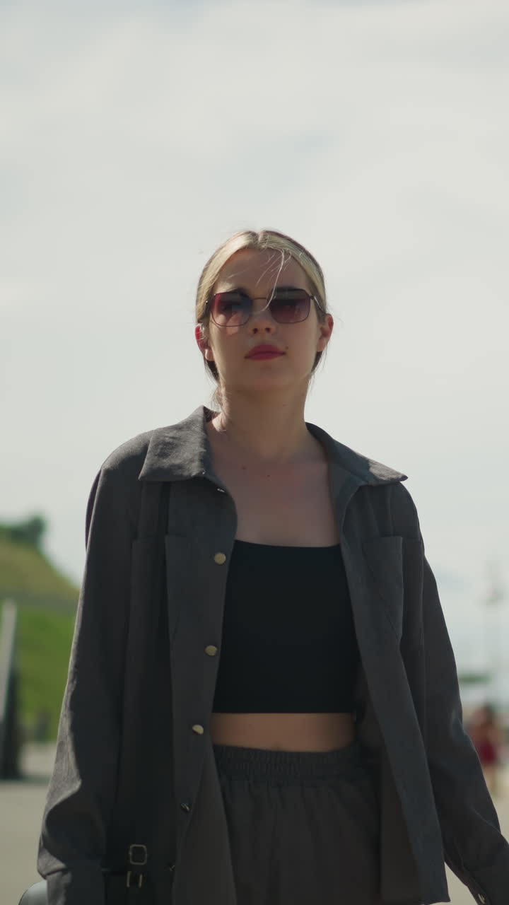 Lady walking confidently on a bridge looks to the right, dressed in a grey shirt over a black crop top and sunglasses, background features blurred view of another person standing in the distance