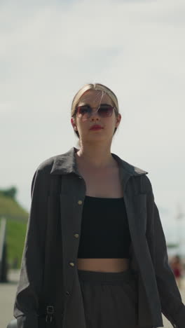 lady walking confidently on a bridge looks to the right, dressed in a grey shirt over a black crop top and sunglasses, background features blurred view of another person standing in the distance
