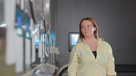 portrait of business owner posing beside industrial washing machine in modern laundromat, stainless drum and blue control screens, confident female in yellow shirt stands for service branding