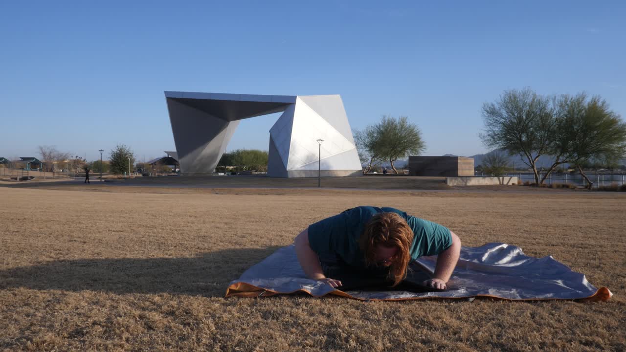 Overweight red hair male doing push ups in a park with an amphitheater behind them.