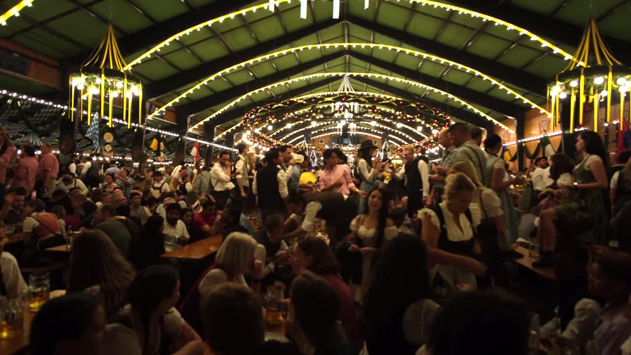 A large crowd in traditional costumes, men in lederhosen and women in dirndls, dance, take selfies, and stand on tables, enjoying Oktoberfest’s lively atmosphere. Everyone is having a great time.