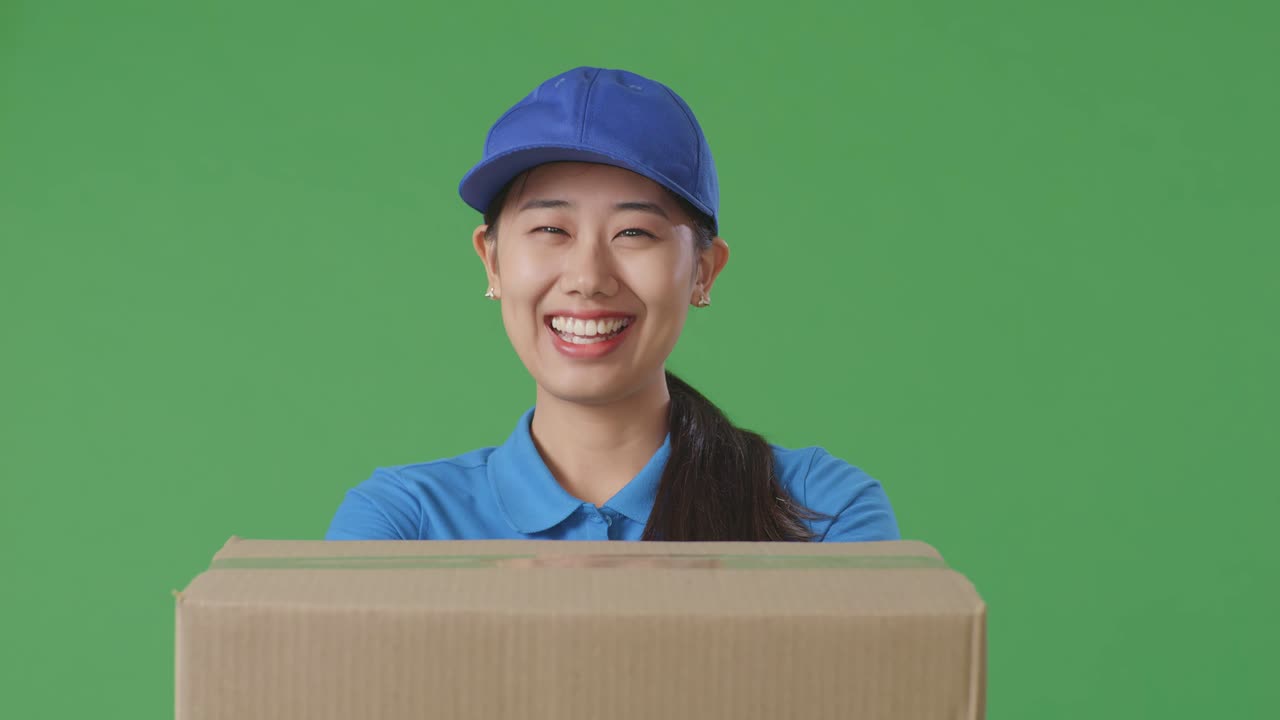 Close Up Of Asian Female Courier In Blue Uniform Showing A Carton To Camera And Smiling While Delivering It On Green Screen Background In The Studio