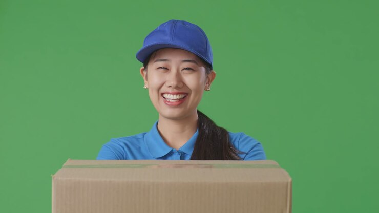 Close Up Of Asian Female Courier In Blue Uniform Showing A Carton To Camera And Smiling While Delivering It On Green Screen Background In The Studio