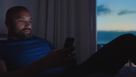 young man using smartphone lying in bed texting browsing social media messages enjoying online entertainment relaxing in apartment at sunset