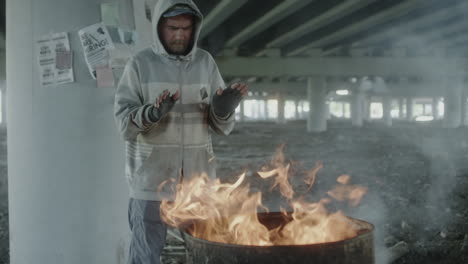 homeless man warming himself by a fire barrel under a bridge