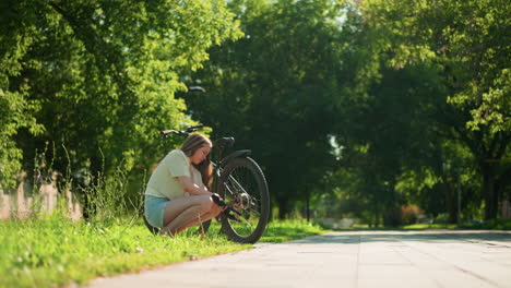 female cyclist crouches under bright sunlight on grassy path, focused on pumping air into back tire of her bicycle, surrounded by lush green trees, with a car passing in the distant background