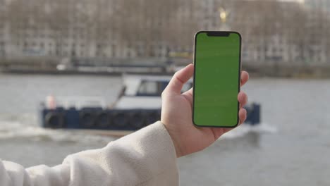 Close-Up-Of-Man-Holding-Green-Screen-Mobile-Phone-Looking-Out-To-River-Thames-And-Embankment-In-London-UK-1