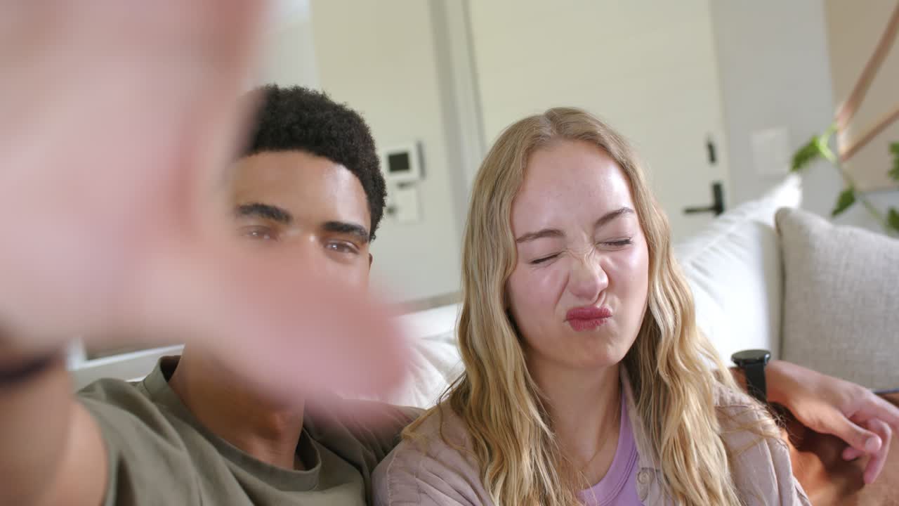 Couple sitting close on couch, making faces while blurred hand entering lens during selfie
