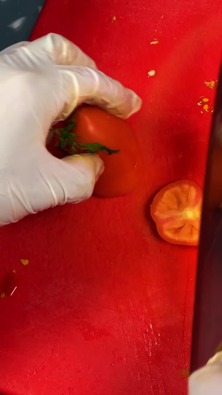 Primo piano delle mani di uno chef con guanti bianchi che affetta un pomodoro rosso. Chef professionista che indossa guanti in lattice taglia un pomodoro fresco su un tagliere di plastica rosso in un ambiente di cucina commerciale.