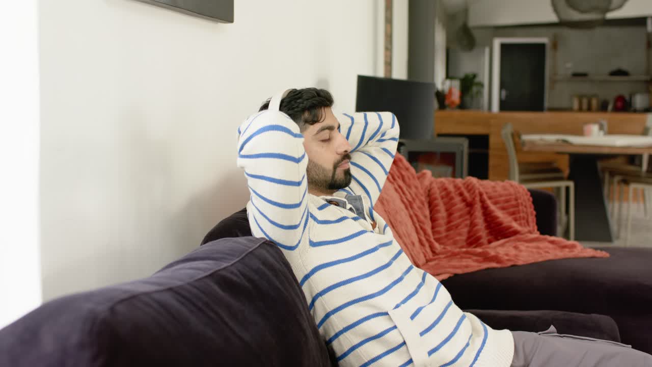 Uomo indiano appoggiato sul divano a casa, con le mani dietro la testa, che si rilassa con un plaid rosso