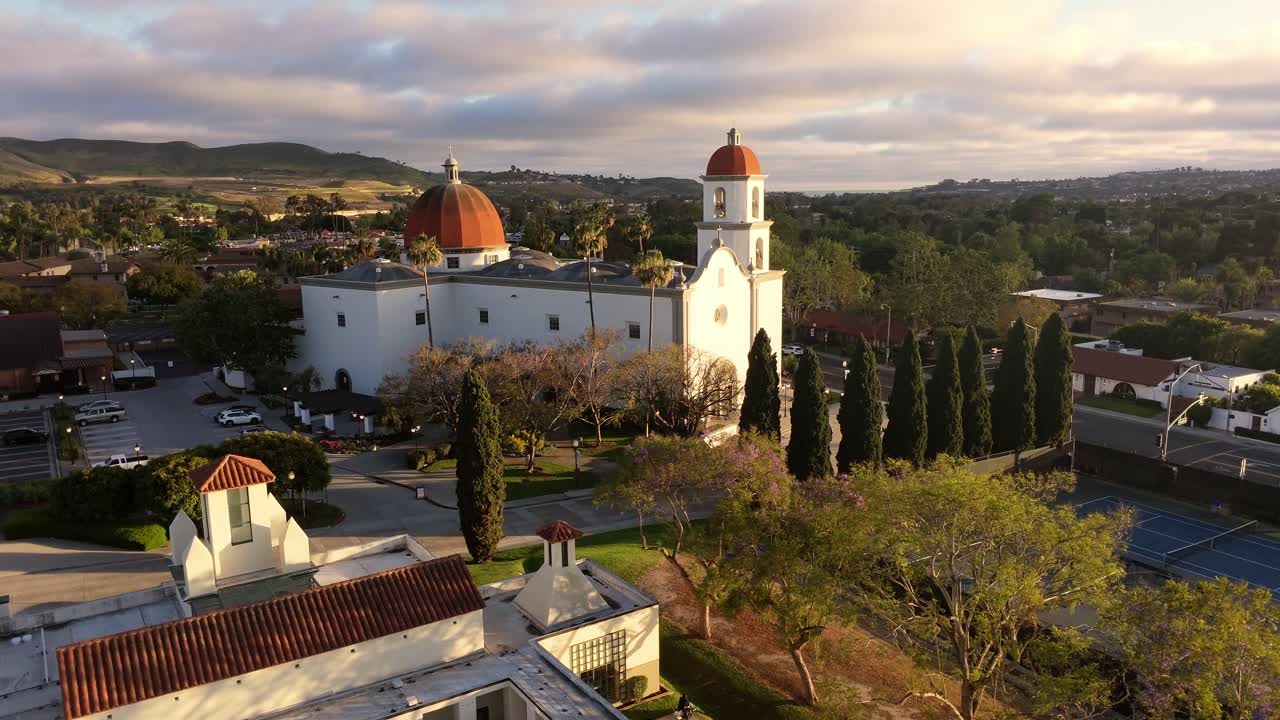 Veduta aerea con drone della Missione Basilica San Juan Capistrano nella calda luce della sera, che mostra la cupola della chiesa, il campanile, i cipressi e le colline circostanti sotto nuvole spettacolari in California