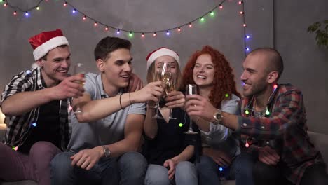 happy young people drinking champagne from the glasses during celebrate new year or christmas eve, having great time in relaxing home atmosphere with garlands lights everywhere. group of young friends some in santa's hats. front view