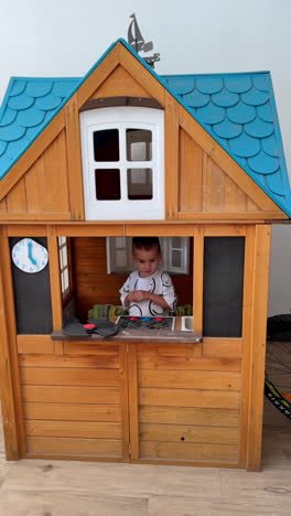 happy caucasian toddler boy plays inside the wooden playhouse. kid leaves the house and peeps from above the corner. top view. vertical video.