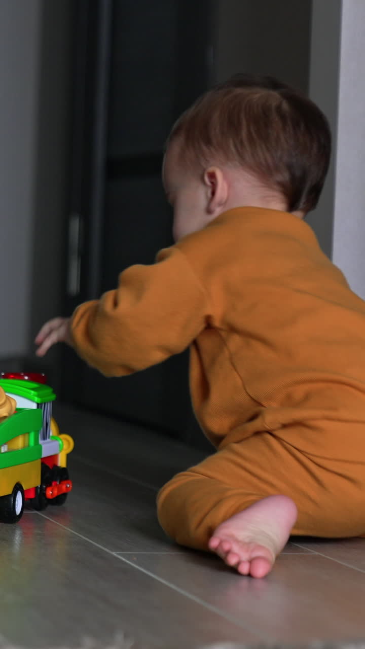 Toy car is moving by the floor and barefoot baby boy follows it. Kid sits on his knees and presses the button on a toy. Rear view. Vertical video