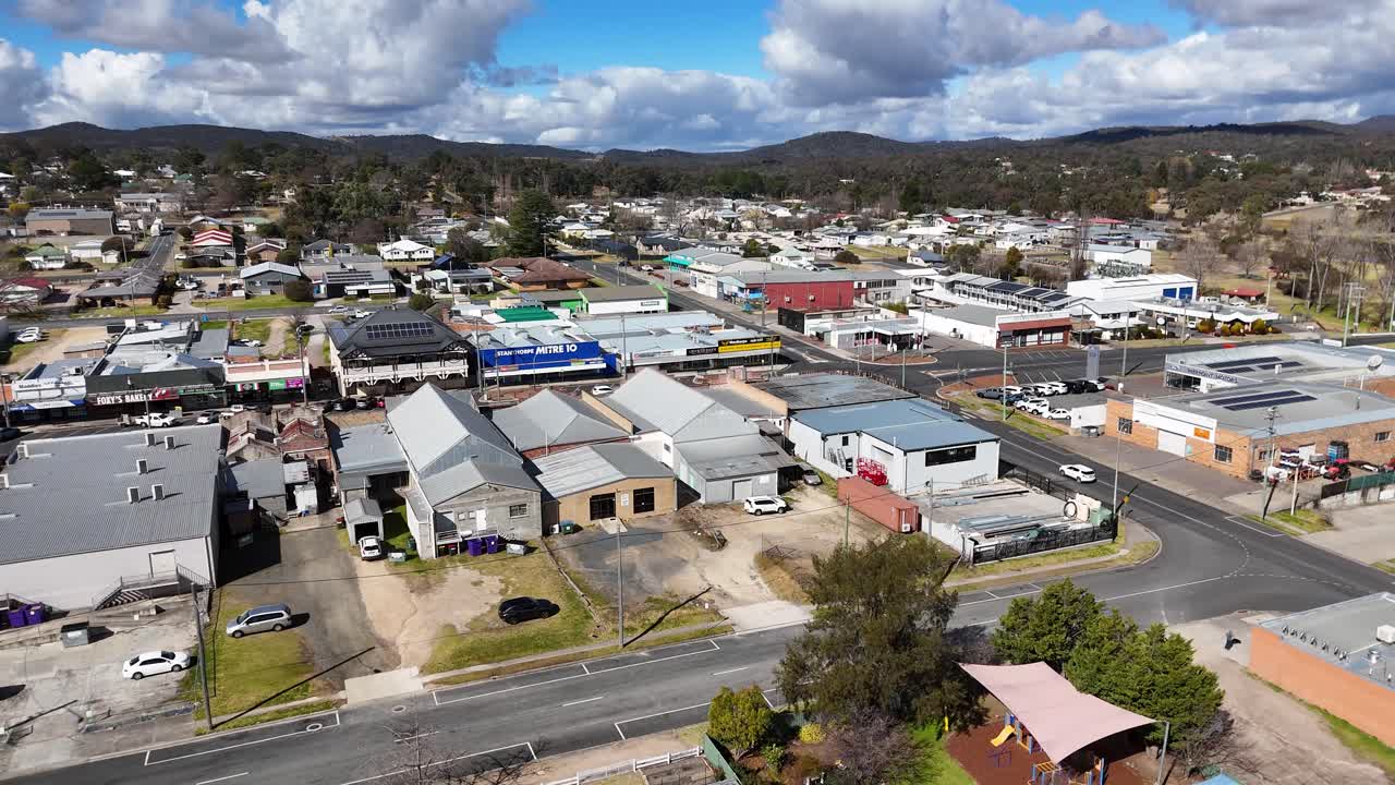 Drone camera smoothly pans above Stanthorpe, Queensland, revealing small-town residential homes, parked cars, and local businesses under bright daylight with scattered clouds