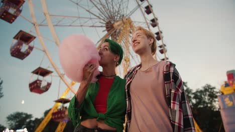 A-blonde-girl-with-a-short-haircut-in-a-pink-plaid-shirt-and-a-girl-with-green-short-hair-in-a-green-shirt-holds-a-huge-pink-cotton-candy-near-the-ferris-wheel-in-the-amusement-park-during-their-date