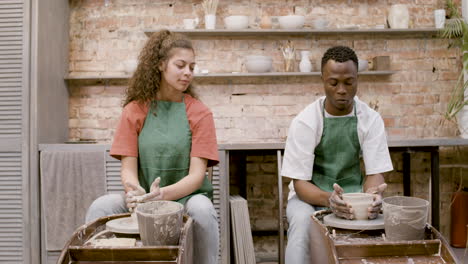 Front-View-Of-Employees-Wearing-Green-Apron-Modeling-Ceramic-Pieces-On-Potter-Wheel-In-A-Workshop-While-Talking-To-Each-Other