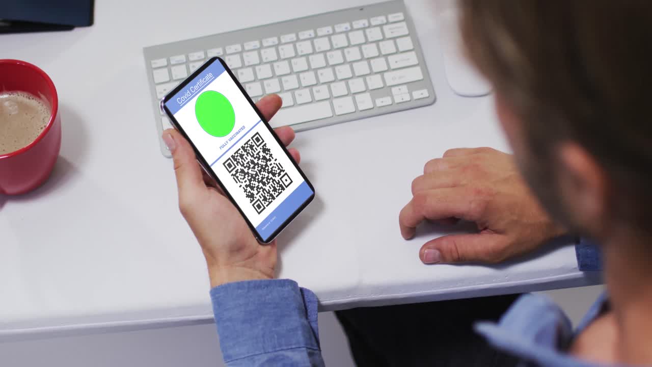 Man at desk holding smartphone with covid vaccination certificate and qr code on screen