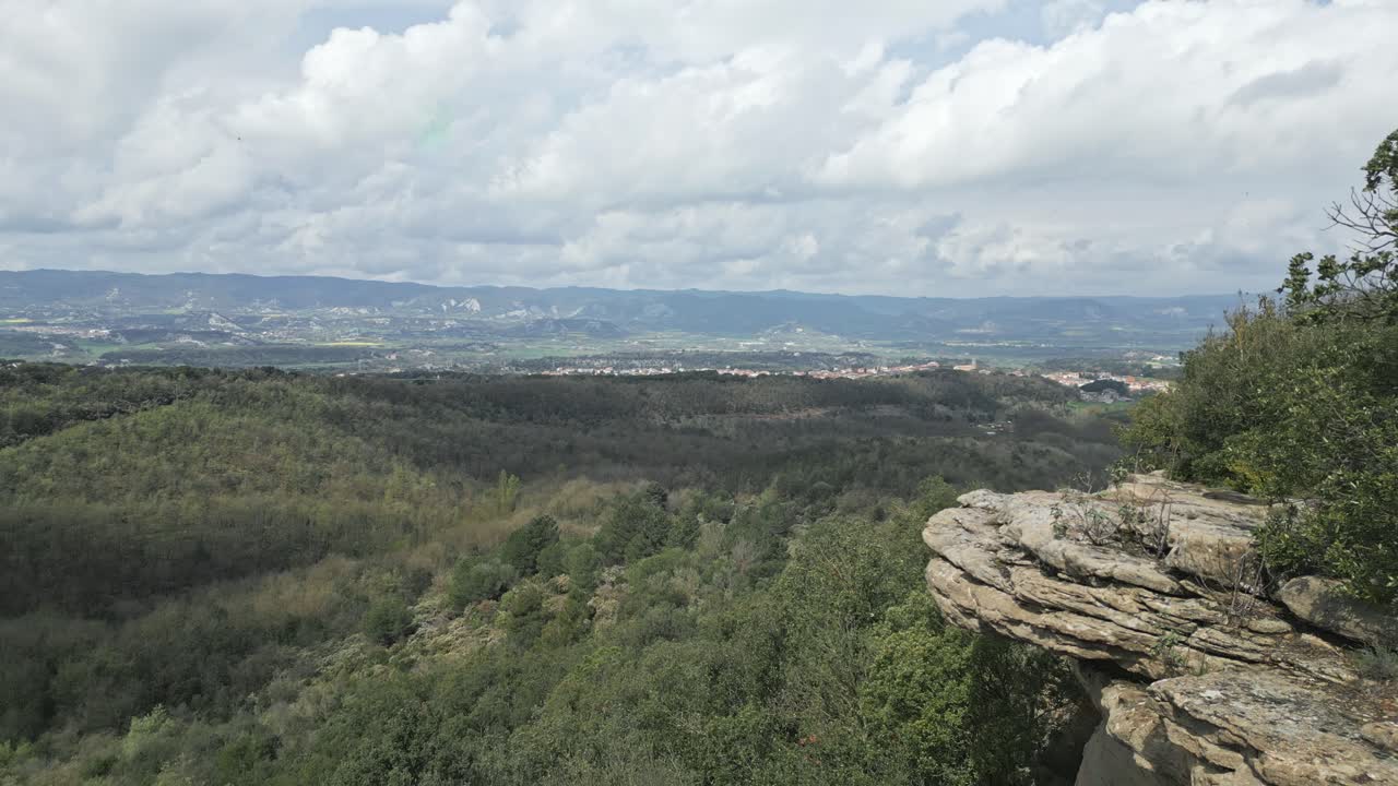Lush green valley stretches out under a cloudy sky, viewed from a rocky cliff edge, showcasing the beauty of untouched nature and distant urban areas