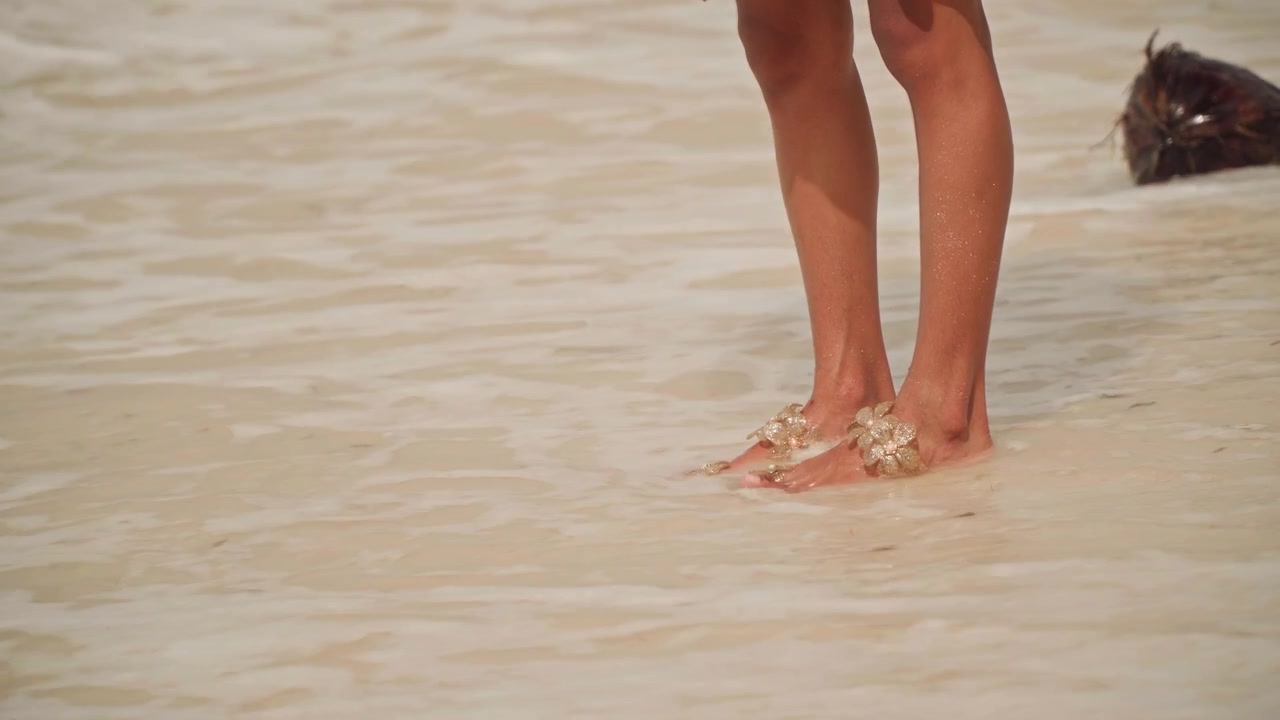 Piedi su sabbia bagnata, bagnasciuga. Primo piano di gambe femminili che indossano sandali decorati mentre camminano tra onde basse, schiuma morbida che lambisce le caviglie, calda luce dorata, impronte sabbiose lasciate indietro.