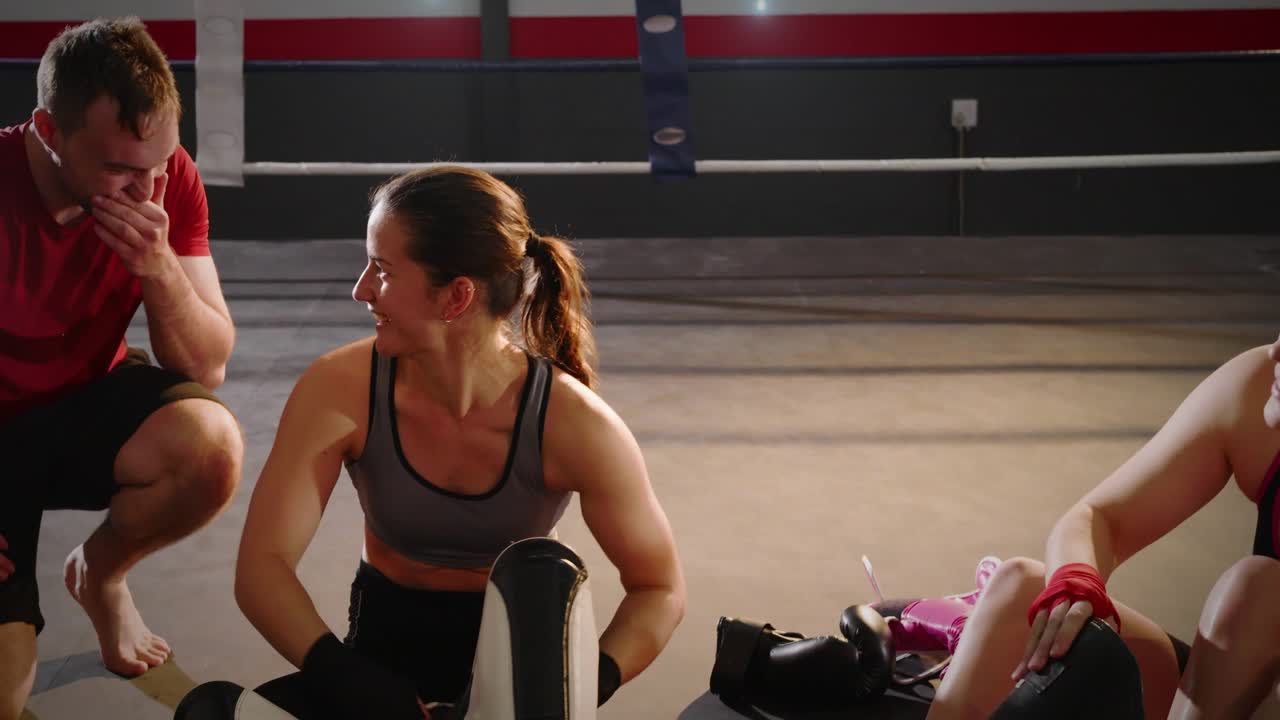 Male walking across ring, kneeling beside seated woman fastening shin guards, team prepping boxing
