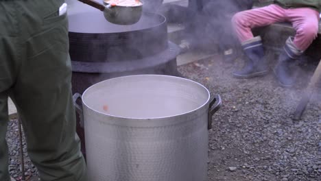 detail of man pouring hot soup into big pot during cold weather with people in back