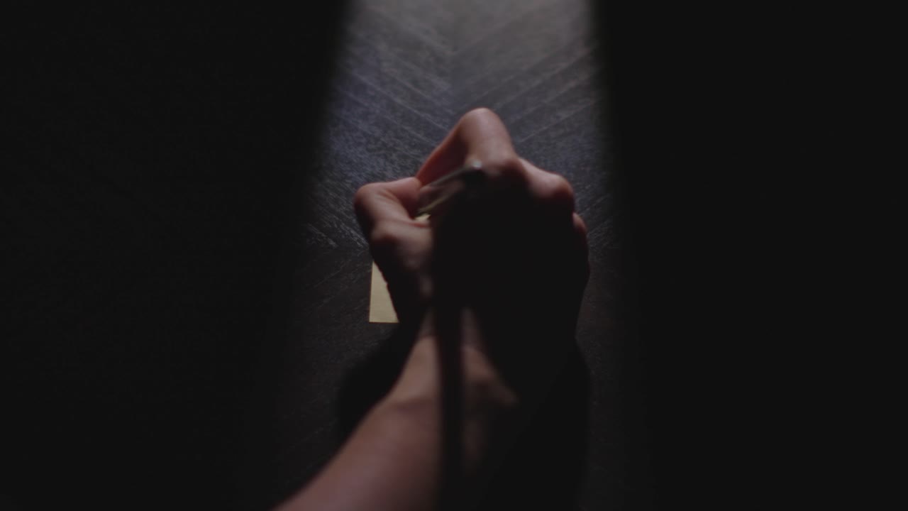 Slow motion close up footage of a hand writing out "This Week" on a paper note and placing it on a table. Shot in dramatic lighting for an impressive title or transition