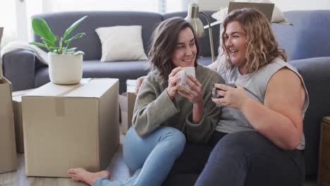 Feliz-Pareja-De-Lesbianas-Caucásicas-Bebiendo-Té-Sentadas-En-El-Suelo-En-Un-Nuevo-Hogar