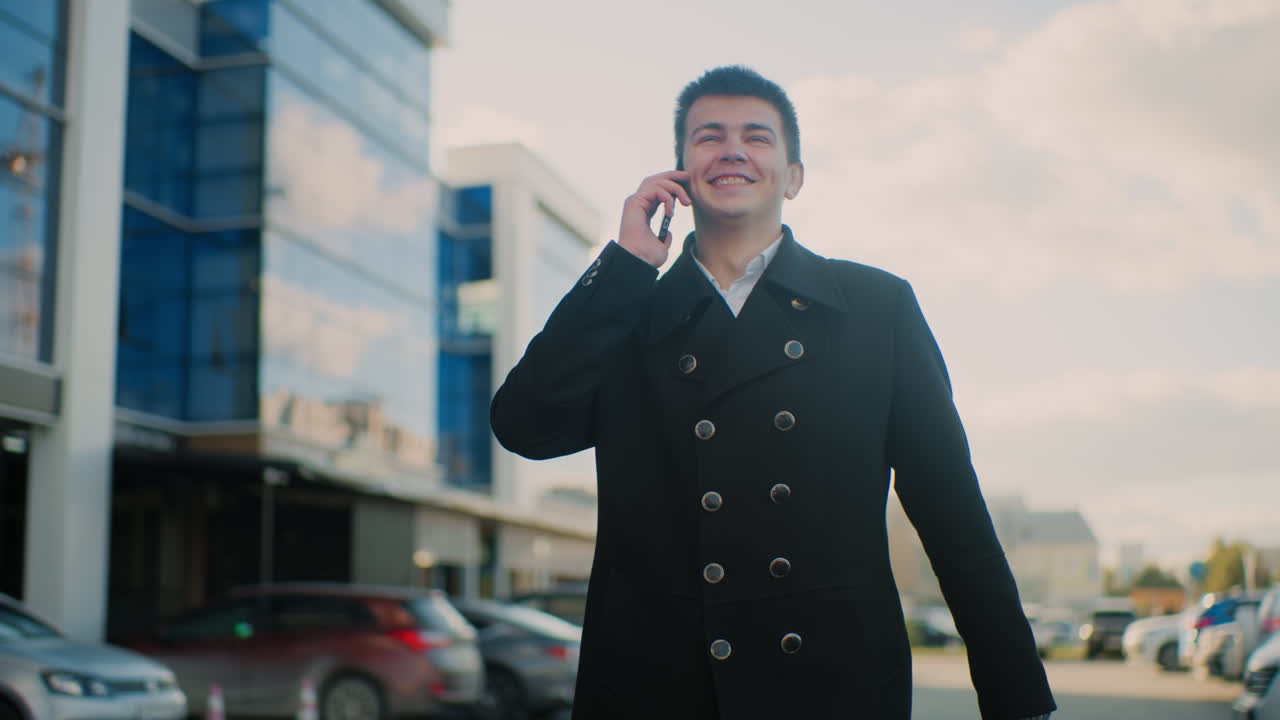 Freelancer in black coat smiling and talking on phone with excitement while gesturing in vibrant urban area featuring modern glass buildings, parked cars, soft sunlight, and cheerful atmosphere