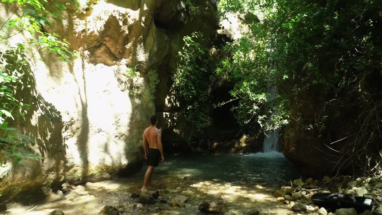 검은색 수영복을 입고 자연림의 신선한 폭포를 바라보며 서있는 백인 젊은 맨발 남자, 뒤로 떠오르는 공중