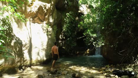 Young-Caucasian-shirtless-and-barefoot-man-wearing-black-bathing-suit-standing-looking-up-at-fresh-waterfall-in-natural-forest,-backward-rising-aerial