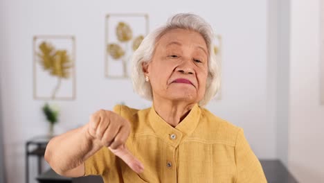 senior asian woman in yellow shirt repeatedly makes thumbs down gesture with serious expression in bright, modern living room. static camera, soft daylight
