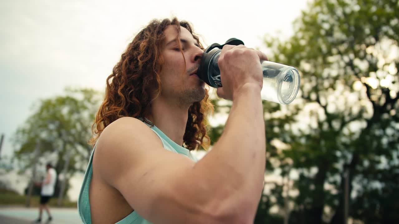 una toma de cerca de un jugador de baloncesto de cabello rojo y pelo rizado en una camiseta de color claro bebiendo agua de una botella deportiva especial, después de lo cual toma la pelota en sus manos y continúa jugando en la cancha de baloncesto en el verano
