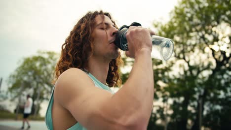 Close-up-shot-of-a-red-haired,-curly-haired-basketball-player-in-a-light-colored-jersey-drinking-water-from-a-special-sports-bottle,-after-which-he-takes-the-ball-in-his-hands-and-continues-to-play-on-the-basketball-court-in-the-summer