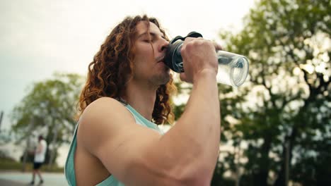 close-up shot of a red-haired, curly-haired basketball player in a light-colored jersey drinking water from a special sports bottle, after which he takes the ball in his hands and continues to play on the basketball court in the summer