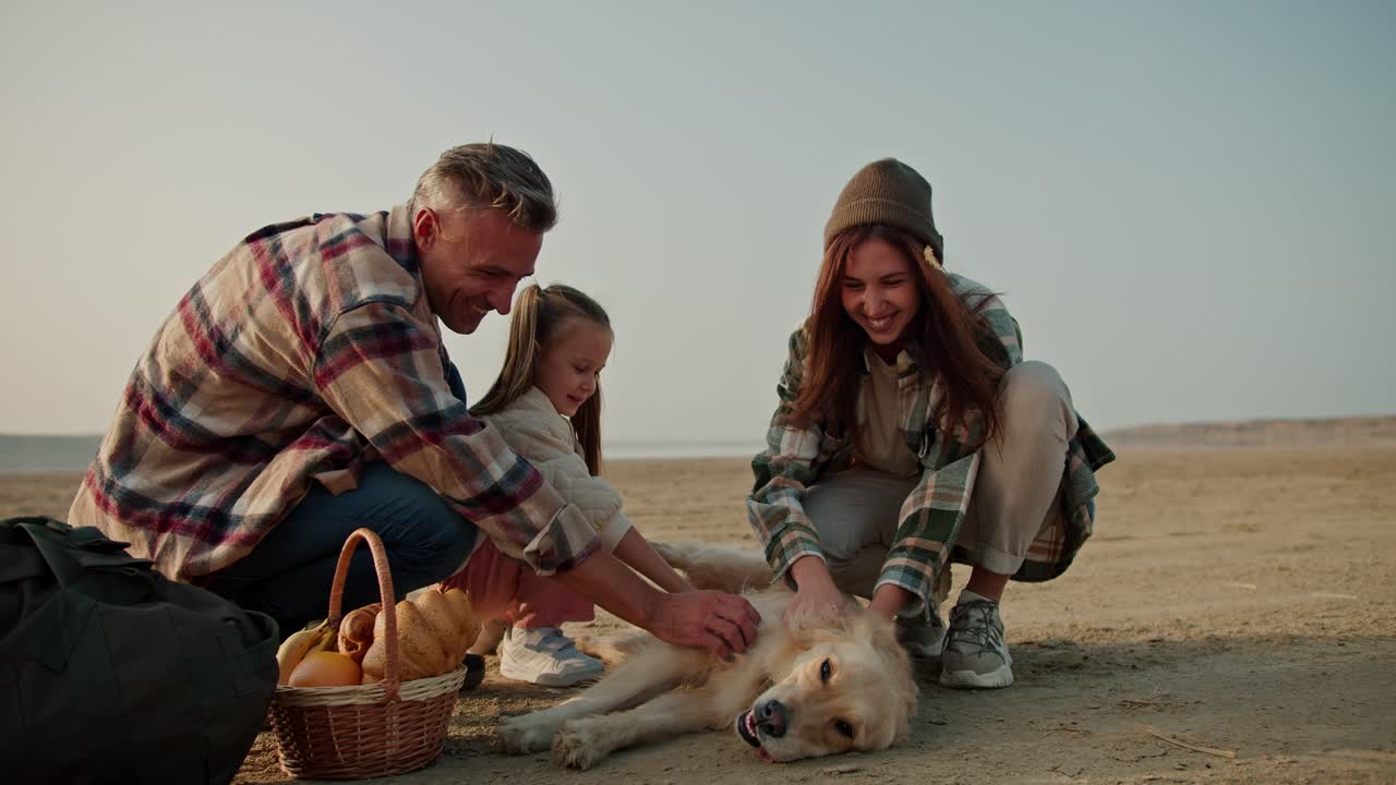 피크닉에서 애완동물 개와 함께 행복한 가족. 아내와 딸과 함께 갈색 체크 셔츠를 입은 행복한 갈색 남자가 여름에 사막 해변에서 피크닉을 하는 동안 큰 크림색 개를 만지고 있습니다.