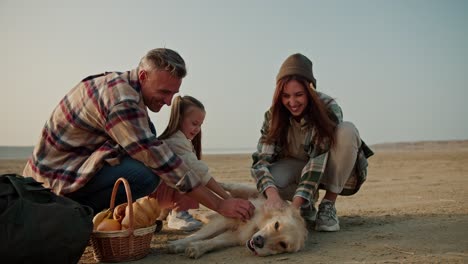 Happy-family-with-their-pet-dog-on-a-picnic.-Happy-brunette-man-in-a-brown-checkered-shirt-with-his-wife-and-daughter-petting-a-large-cream-colored-dog-during-their-picnic-on-a-deserted-seashore-in-summer