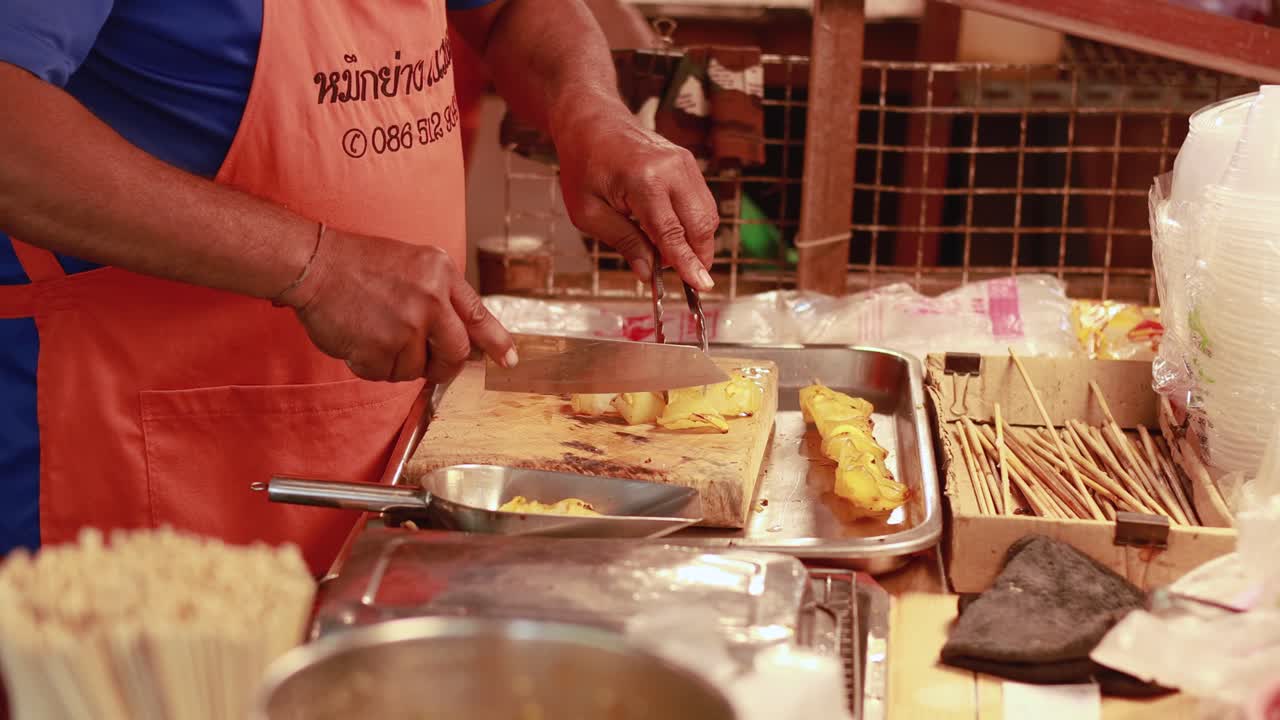 A vendor skillfully slices food with a knife and tongs at a bustling Bangkok floating market. Warm lighting enhances the vibrant scene